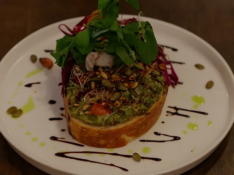 Avocado smash on toast piled with sprouts, seeds and shredded beetroot, on a white plate with a balsamic drizzle.