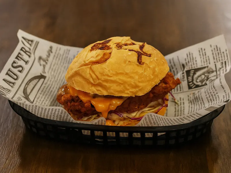 A fried chicken burger with melted cheese and slaw in a golden bun, served in a black basket lined with newsprint paper.