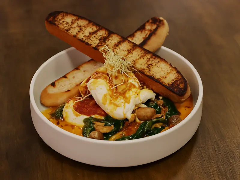 Poached eggs in a white bowl over spinach and chilli oil, with two grilled bread batons resting across the rim.